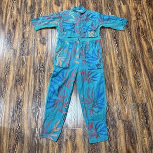 May include: A teal jumpsuit with a button-up top and long trousers. The jumpsuit features a colorful leaf pattern in red, blue, and orange. It has multiple pockets and short sleeves. The garment is laid flat on a wooden surface.