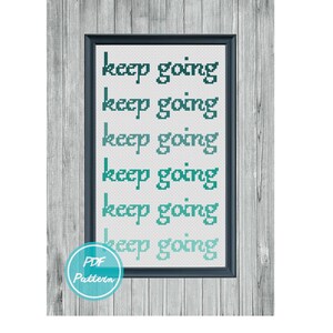 Puede incluir: Un patrón de punto de cruz con la frase "keep going" repetida seis veces en un degradado de colores turquesa y verde. El patrón está enmarcado en un marco negro y se muestra sobre un fondo de madera.