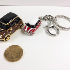 Puede incluir: Dos llaveros de coche en miniatura, uno marr&oacute;n con techo negro y otro rojo con techo blanco, unidos a un llavero plateado.