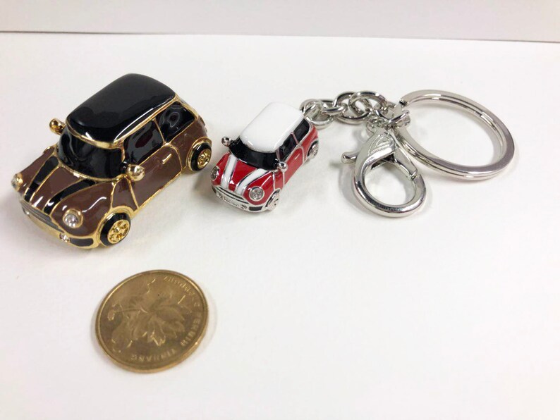 Puede incluir: Dos llaveros de coche en miniatura, uno marr&oacute;n con techo negro y otro rojo con techo blanco, unidos a un llavero plateado.