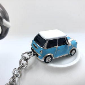 May include: A small, light blue car charm with a white roof and black windows, connected to a silver chain. The car has silver rims and a side mirror. The keychain is set against a white background.