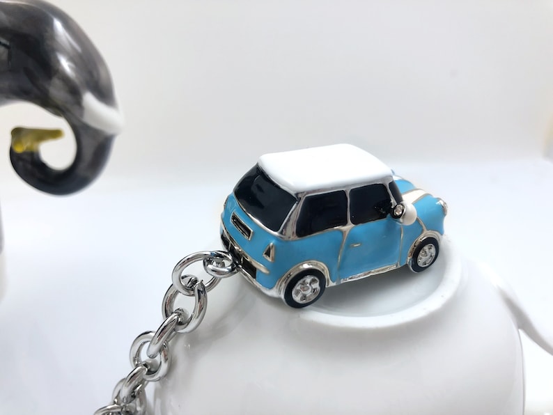 May include: A small, light blue car charm with a white roof and black windows, connected to a silver chain. The car has silver rims and a side mirror. The keychain is set against a white background.