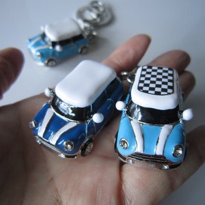 May include: Miniature car keyrings in blue, white, and silver. The keyrings feature a classic car design with a checkered roof on one. The cars have a glossy finish and are shown in a hand.