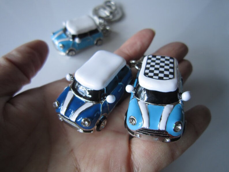 May include: Miniature car keyrings in blue, white, and silver. The keyrings feature a classic car design with a checkered roof on one. The cars have a glossy finish and are shown in a hand.