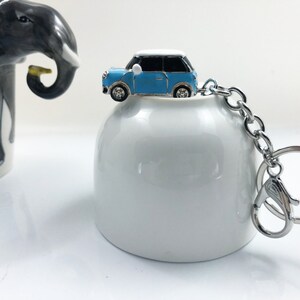 May include: A miniature blue and white car keyring with a silver chain and clasp. The car has black windows and silver wheels. The keyring is on a white ceramic object. An elephant-themed mug is in the background.