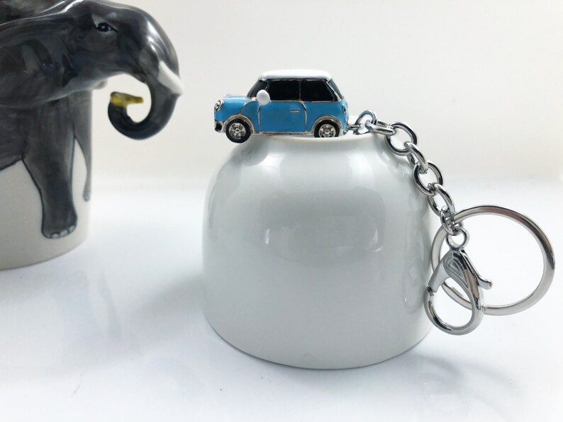 May include: A miniature blue and white car keyring with a silver chain and clasp. The car has black windows and silver wheels. The keyring is on a white ceramic object. An elephant-themed mug is in the background.