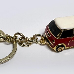 Puede incluir: Un llavero dorado con un coche en miniatura rojo y blanco con techo blanco. El coche tiene un interior negro y ruedas doradas.