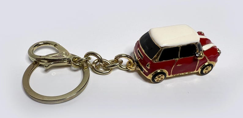 Puede incluir: Un llavero dorado con un coche en miniatura rojo y blanco con techo blanco. El coche tiene un interior negro y ruedas doradas.
