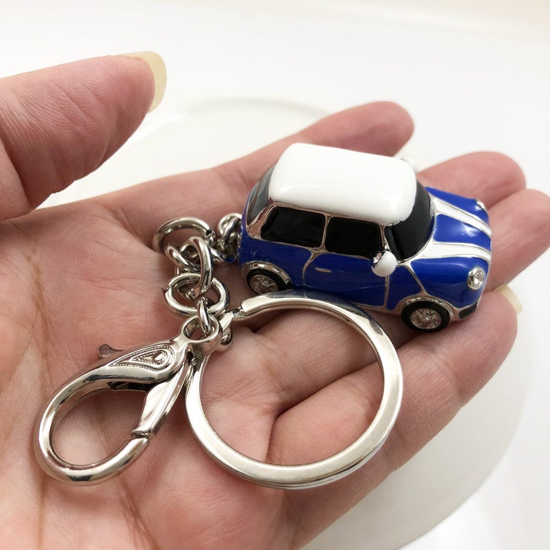 May include: A blue and white miniature car keyring with a silver ring and clasp. The car has a white roof and two white stripes on the bonnet. The keyring is held in a hand.