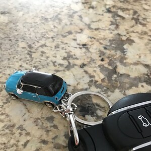 May include: A blue and black car-shaped keyring with a silver ring and clasp. The keyring is next to a black car key fob with white icons. The car keyring is a miniature replica of a car.