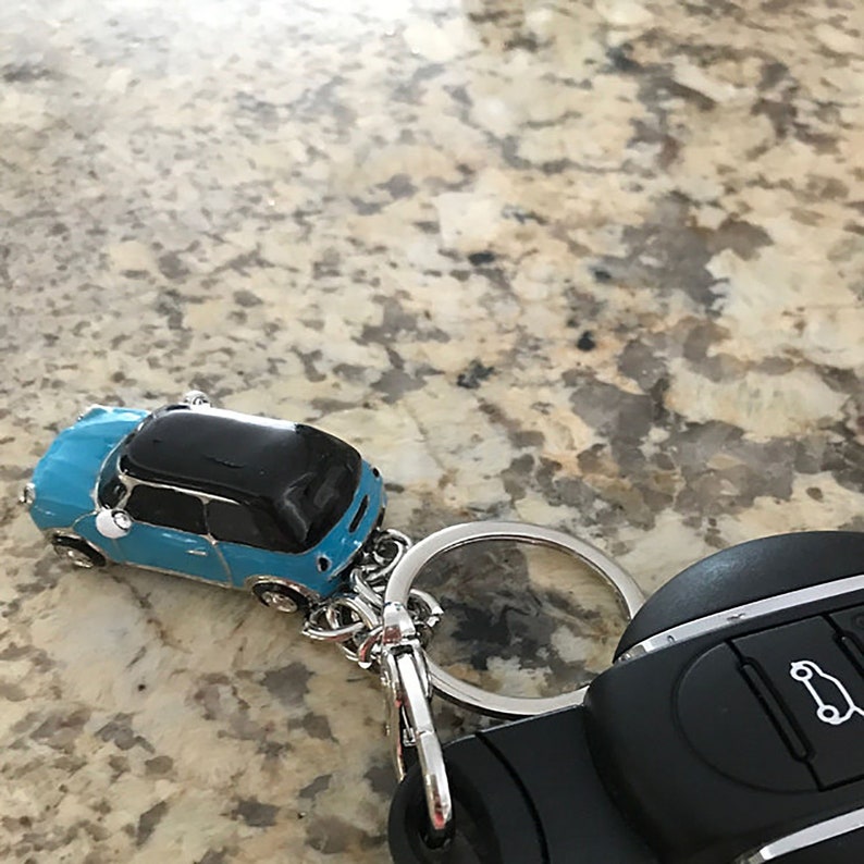 May include: A blue and black car-shaped keyring with a silver ring and clasp. The keyring is next to a black car key fob with white icons. The car keyring is a miniature replica of a car.