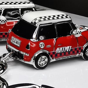 May include: Two red and white Mini Cooper car keychains with silver accents. The cars have black checkered patterns on the roof and sides, with the word "MINI" displayed. The keychains are on a white surface.