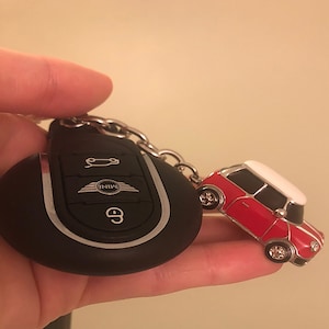 May include: Black car key fob with silver trim and a red and white Mini Cooper car keyring. The keyring car has silver wheels and a white roof. The key fob has the Mini logo and a button to unlock the car.