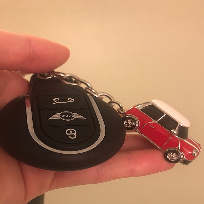 May include: Black car key fob with silver trim and a red and white Mini Cooper car keyring. The keyring car has silver wheels and a white roof. The key fob has the Mini logo and a button to unlock the car.