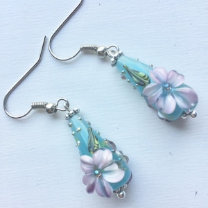 Peut inclure: Une paire de boucles d'oreilles pendantes avec des perles de verre en forme de larme bleu clair. Chaque perle est décorée d'une fleur rose et d'accents argentés.