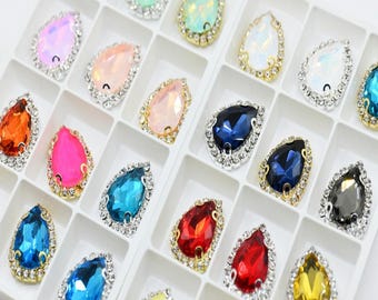 May include: A tray of colorful teardrop-shaped rhinestones. Each rhinestone is set in a metal frame and surrounded by smaller, clear crystals. The rhinestones come in various colors, including pink, blue, orange, and yellow.