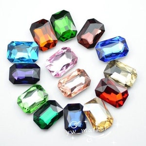 Puede incluir: Surtido de 14 piedras de cristal talla esmeralda en varios colores, incluyendo azul, verde, rojo, morado, rosa y dorado. Las piedras están dispuestas en un patrón circular sobre un fondo blanco.
