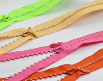 Puede incluir: Cuatro cremalleras de diferentes colores con adornos de strass transparentes. Las cremalleras son de color verde, beige, rosa y naranja.