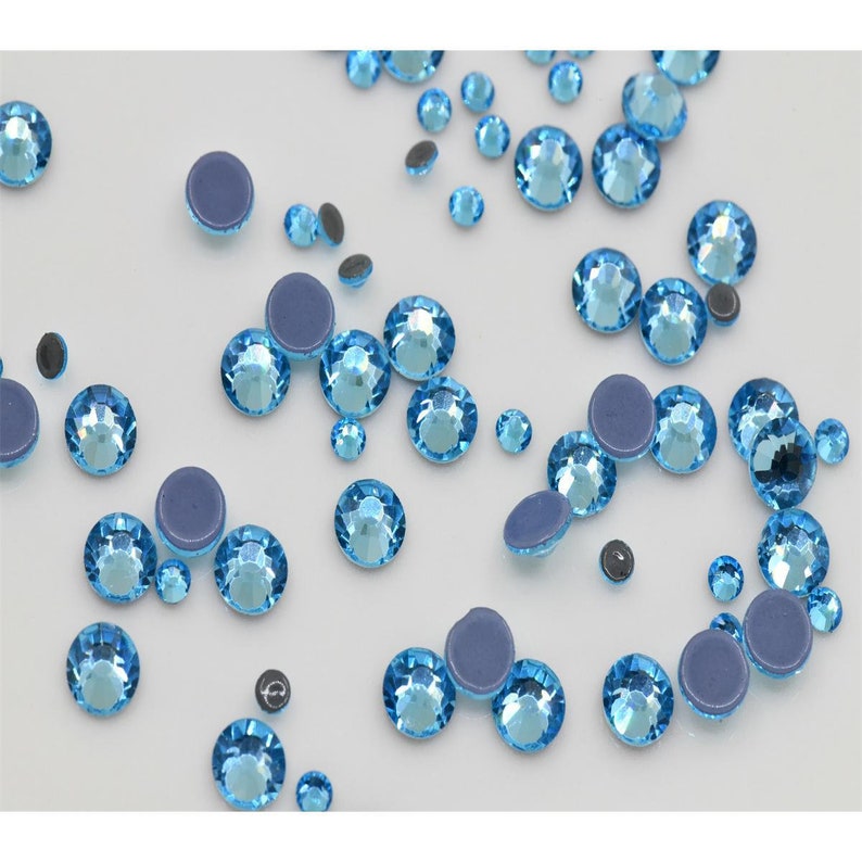 May include: Assortment of light blue round rhinestones with a flat back. The rhinestones are scattered on a white surface.