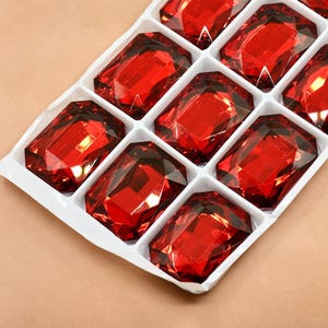 Op de afbeelding: Een close-up van een verpakking met rode, gefacetteerde, rechthoekige strasssteentjes. De strasssteentjes zijn gerangschikt in een rasterpatroon in een doorzichtige plastic container.