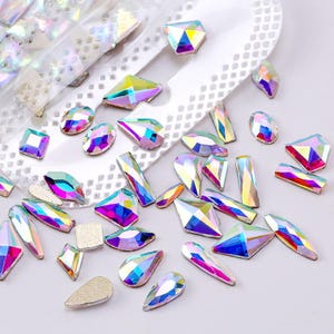 May include: A collection of iridescent rhinestones in diverse shapes, such as squares, ovals, and teardrops. The rhinestones display a multi-coloured, shimmering effect, reflecting shades of pink, blue, and yellow. Some are on a clear adhesive sheet.