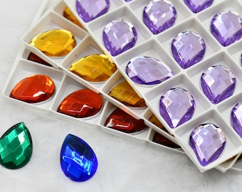 May include: Assortment of colorful teardrop-shaped rhinestones in a plastic storage container. The rhinestones are arranged in rows and are available in purple, orange, red, and green.