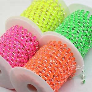 Puede incluir: Cuatro carretes de ribete de cadena de strass de color neón. Los carretes están enrollados con cadena plateada y strass de color rosa neón, naranja, amarillo y verde.