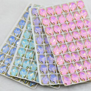 May include: Three trays of iridescent glass rhinestones in shades of light blue, lavender, and pink. The rhinestones are arranged in a grid pattern and have a pear shape.