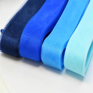 Könnte beinhalten: Vier Rollen blauen Tüllstoffs in verschiedenen Farbtönen: Marineblau, Königsblau, Mittelblau und Hellblau. Der Tüll ist eng gewebt und wirkt weich. Die Rollen liegen nebeneinander, die Kanten des Stoffes sind sichtbar.