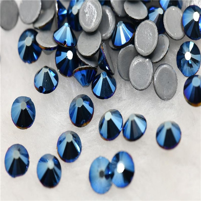 May include: A scattering of dark blue iridescent rhinestones on a white background. Some of the rhinestones are loose, while others are attached to silver backing.