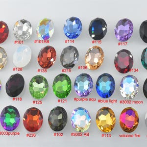 May include: A collection of oval-shaped faceted crystals arranged in four rows. Each crystal features a unique color or finish, including red, clear, iridescent, blue, black, purple, gold, and green. Each stone is labeled with a red number or name.