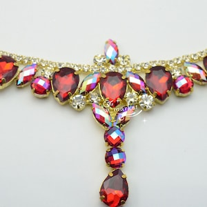May include: A gold-toned decorative piece with a series of red, teardrop-shaped rhinestones. The piece also features smaller, iridescent and clear rhinestones. The design is symmetrical, with a central pendant of red rhinestones.