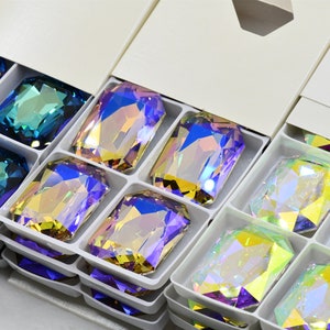 May include: Assortment of iridescent glass gems in various colours, including blue, green, purple, and yellow, arranged in a white box with compartments. The gems are rectangular and have a faceted cut.