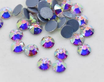 May include: Assortment of iridescent clear glass rhinestones with a purple hue. The rhinestones are scattered on a white surface.