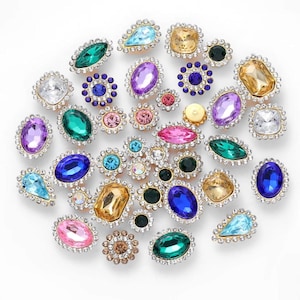 May include: Assortment of colourful rhinestone embellishments in various shapes and sizes, including round, oval, teardrop, and flower designs. The rhinestones are set in gold metal settings.