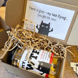 Puede incluir: Una caja de cart&oacute;n marr&oacute;n llena de papel triturado y un frasco transparente etiquetado MY LITTLE WTF JAR con 60 afirmaciones. La tapa interior presenta una tarjeta blanca con una ilustraci&oacute;n de un gato negro y el texto: There it goes&mdash;my last flying... (you get the idea).