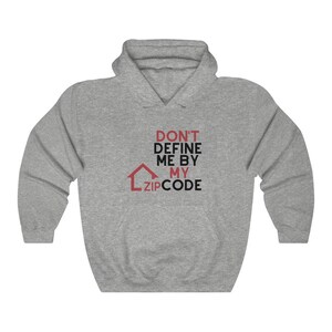Op de afbeelding: Een grijze hoodie met de tekst "DON'T DEFINE ME BY MY ZIP CODE" in rood en zwart. Een rode huisafbeelding staat onder de tekst. De hoodie heeft een capuchon met trekkoord en een voorzak.