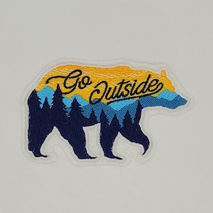 May include: Embroidered patch in the shape of a bear, featuring a landscape design with the text "Go Outside". The patch has a yellow and blue sky, blue trees, and a dark blue bear silhouette.