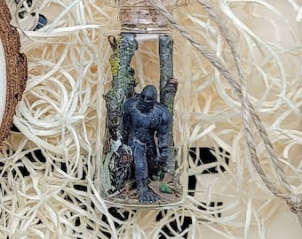May include: A glass bottle ornament with a string for hanging. Inside the bottle is a small figurine of a creature resembling Bigfoot standing in a forest setting.
