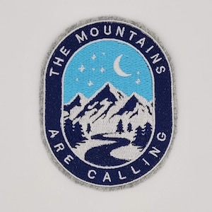 May include: Embroidered patch with a mountain landscape design. The patch features a blue and white mountain scene with a crescent moon and stars against a light blue sky. The text "THE MOUNTAINS ARE CALLING" is in a navy blue border.