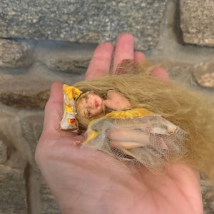 Puede incluir: Una pequeña muñeca hecha a mano con cabello largo y rubio, vestida con un vestido amarillo y blanco con estampado floral. La muñeca está acostada de lado con los ojos cerrados, como si estuviera durmiendo.