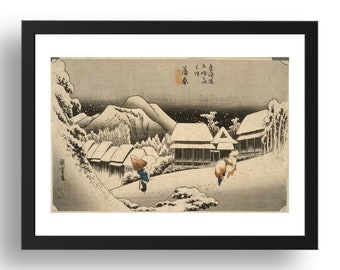 Op de afbeelding: Een ingelijste Japanse houtsnede met een besneeuwd landschap met traditionele huizen, twee figuren die over een pad lopen en bergen op de achtergrond. Het kunstwerk bevat gedempte tinten grijs, wit en blauw met Japanse kalligrafie.