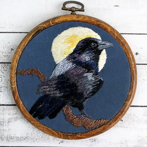 May include: Embroidered hoop art featuring a black crow with a yellow moon in the background. The crow is perched on a branch with a blue background.