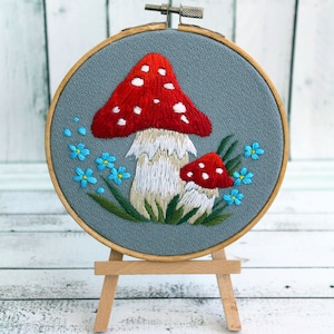 May include: Embroidered hoop art featuring two red and white mushrooms with white stems and blue forget-me-not flowers on a gray background.