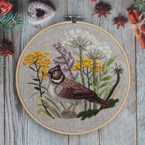 May include: Embroidered hoop art featuring a brown and white bird perched on a branch with green leaves and yellow flowers. The background is a beige linen fabric.