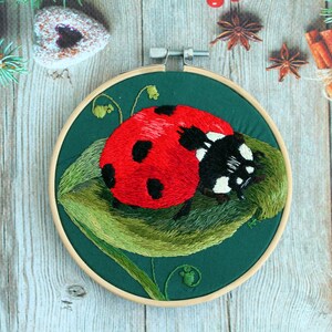 May include: Embroidered ladybug on a green leaf, framed in a wooden hoop. The ladybug is red with black spots and white accents.