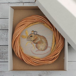 May include: A brown wicker wreath with a beige fabric center featuring an embroidered image of a brown mouse holding a stalk of wheat.