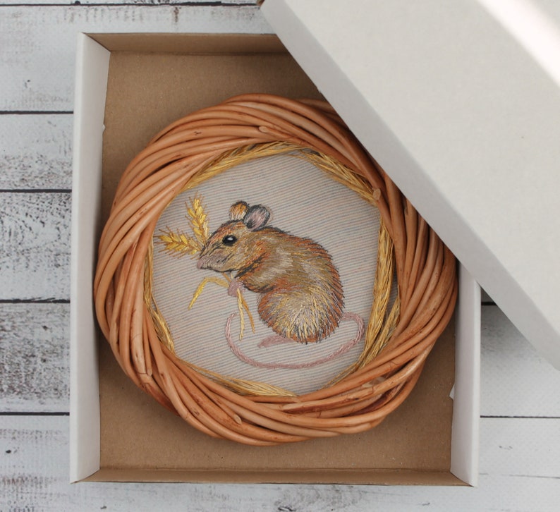 May include: A brown wicker circle with a beige fabric center featuring an embroidered image of a brown mouse holding a stalk of wheat.