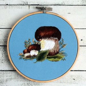 May include: Embroidered hoop art featuring three mushrooms with brown caps and white stems, surrounded by green leaves and yellow flowers on a blue background.