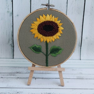May include: Embroidered sunflower with brown and black seed details on a gray fabric stretched over a wooden embroidery hoop. The hoop is mounted on a wooden easel.